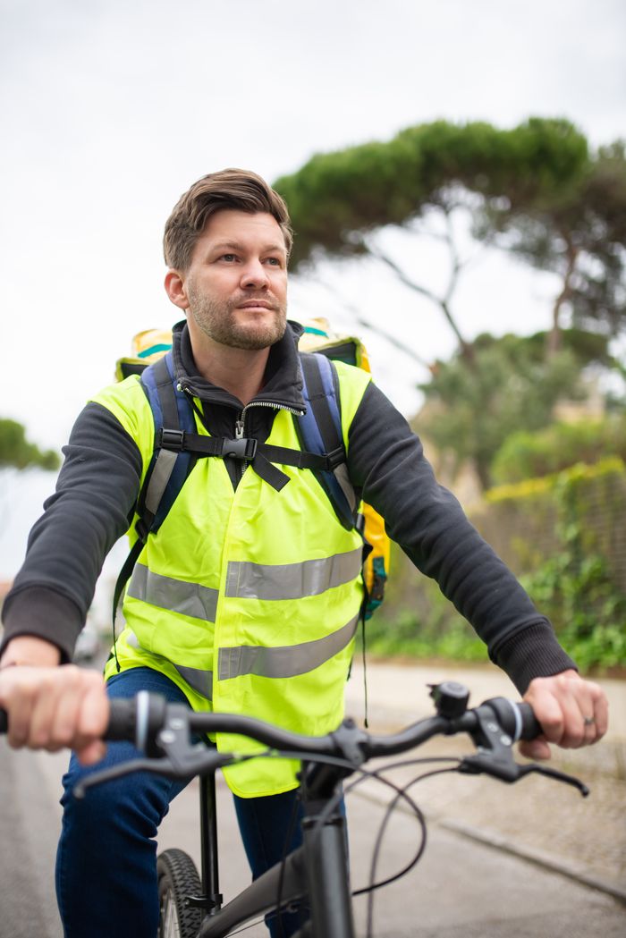 A male courier riding a bicycle for delivery service, wearing a reflective vest. Outdoor scene.